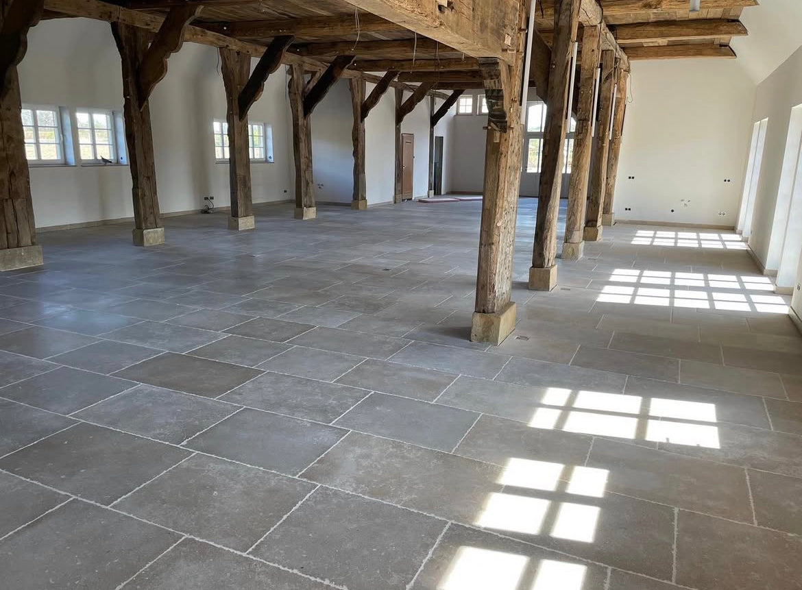 Large room with wooden beams and stone flooring, sunlight streaming through windows.
